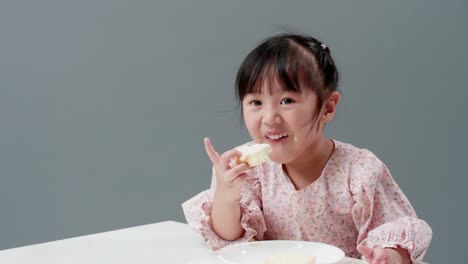 Asiatisches-Kind,-Das-Im-Studio-Ein-Stück-Süßes-Mit-Grauem-Hintergrund-Isst