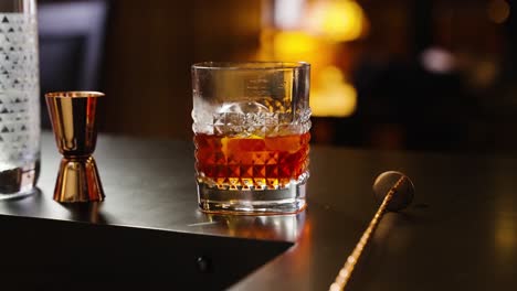 Negroni-Cocktail-with-spherical-ice-back-lit-on-top-of-a-bar-with-jigger-and-mixing-stick