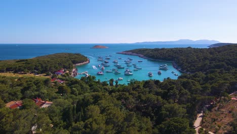 Yachts-in-the-bay-near-the-coastal-city-of-Croatia-against-the-backdrop-of-blue-skies-and-blue-transparent-water,-green-lush-trees-and-houses-with-red-roofs