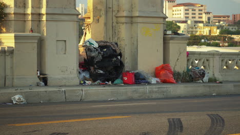 View-of-bags-full-of-trash-piled-up-on-the-sidewalk-of-6th-st-bridge,California,-USA-with-cars-passing-by-in-high-speed