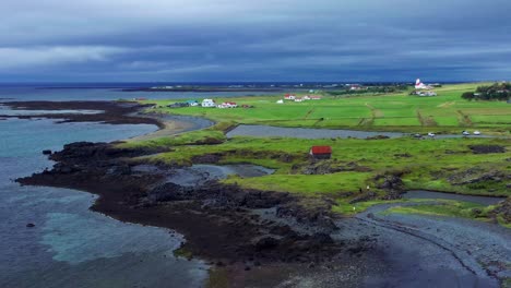 Aerial-View-Of-Volcanic-Coastline-With-Green-Fields-And-Houses-In-Gardakirkjugardur,-Gardabaer,-Iceland