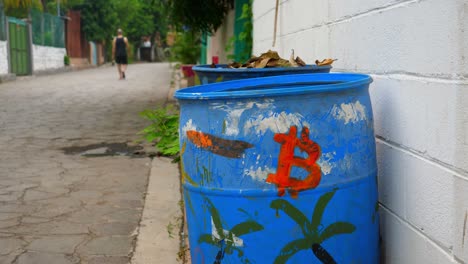 Full-shot,-a-blue-drum-with-a-red-logo-of-bitcoin-on-the-bitcoin-beach-in-El-Salvador,-Mexico,-a-person-walking-in-the-background