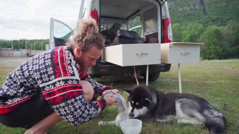 Männlicher-Camper-Gießt-Hundefutter-In-Eine-Plastikschüssel-Für-Sein-Haustier-Alaskan-Malamute