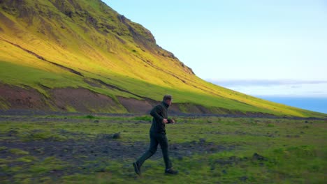 Caucasian-Man-Running-On-Grassy-Terrain-Near-The-Mountains-Of-Seljavallalaug-In-Southern-Iceland