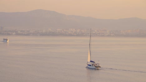 Sunrise,-sunset-over-Adriatic-with-yacht-floating-in-the-sea-and-coastal-city-in-the-background