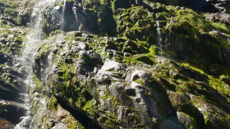Tilt-down-shot-of-waterfall-cascading-rocky-mountain-with-moss-during-sunny-day---Fiordland-National-Park,New-Zealand