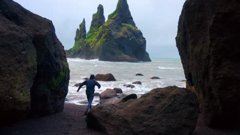 Man-Running-Through-Boulders-At-Reynisfjara-Black-Sand-Beach-In-Iceland-With-View-Of-Reynisdrangar-Basalt-Rock-Formation-In-Background