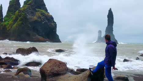 Male-Tourist-At-The-Reynisfjara-Black-Sand-Beach-In-Iceland,-Raising-Hands-As-Waves-Crash-On-The-Rocks