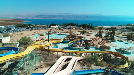 drone-shot-of-a-Couple-sitting-on-a-water-slide-in-an-abandoned-park,-in-front-of-the-dead-sea