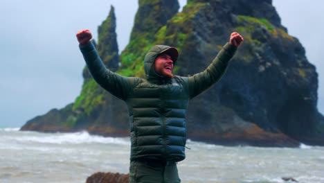 Happy-Male-Tourist-In-Winter-Jacket-With-Hood-Raising-Arms-Standing-On-Coastal-Rock-At-Reynisfjara,-Southern-Iceland