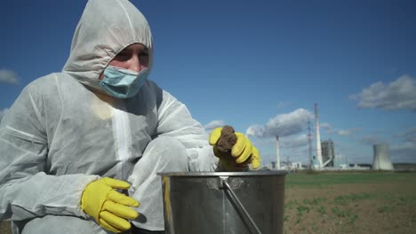 Scientist-in-protective-suit-checking-contamination-of-field-clay,-industrial-background,-ecological-disaster-concept