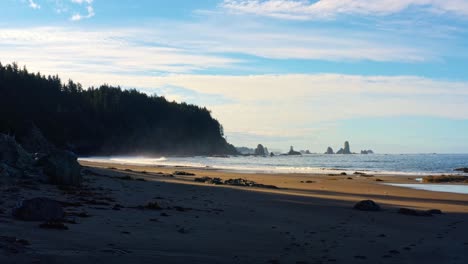Stunning-drone-time-lapse-of-the-gorgeous-Third-Beach-in-Forks,-Washington-with-large-rock-formations,-surrounded-by-a-pine-tree-forest-on-cliffs,-and-golden-sand-on-a-warm-summer-morning