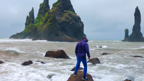 Man-In-Blue-Jacket-Standing-On-A-Rock-Keeping-His-Balance-At-Reynisfjara-Black-Sand-Beach-In-Iceland-On-A-Windy-Day