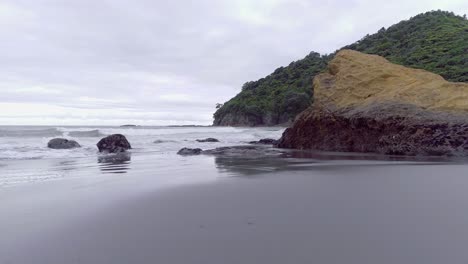 Flying-low-past-rocky-jagged-cliffs-on-beach-in-New-Zealand,-cloudy-day
