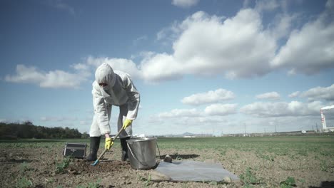 Scientist-in-protective-suit-digging-contaminated-field-clay,-industrial-background,-ecological-catastrophe-disaster-concept
