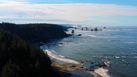 Stunning-aerial-drone-shot-of-the-gorgeous-Third-Beach-in-Forks,-Washington-with-large-rock-formations,-surrounded-by-a-pine-tree-forest-on-cliffs,-and-golden-sand-on-a-warm-summer-morning