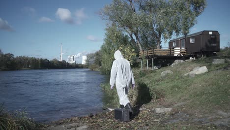 Woman-in-protective-suit-walking-towards-river-to-do-water-sample,-holding-test-tube-and-case,-follow-dolly-shot