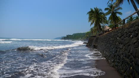 Full-shot,-wave-hitting-the-walls-of-the-bitcoin-beach-on-a-bright-Sunday-in-El-Salvador,-Mexico,-palm-trees-and-bright-blue-sky-in-the-background