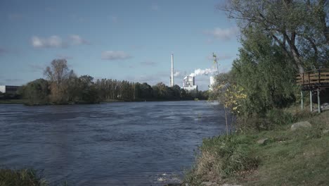 Woman-in-protective-suit-walking-towards-camera,-river-and-smoking-factory-in-background,-outdoor-middle-shot-view