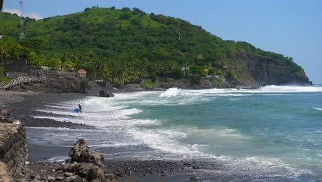 Full-shot,-a-surfer-walks-out-the-beach-on-a-bright-sunny-day-at-the-bitcoin-beach-in-El-Salvador,-Mexico,-scenic-view-of-the-beach-and-cliff-in-the-background