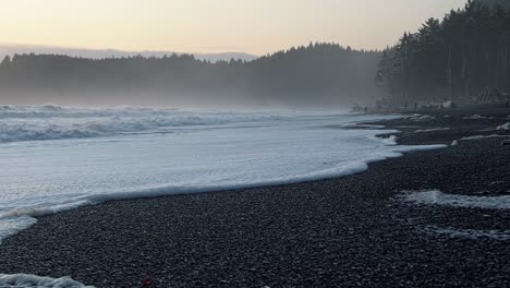 Tilting-up-shot-of-the-coastline-of-the-famous-Ruby-Beach-near-Forks,-Washington-with-small-waves-crashing-and-leaving-sea-foam-behind,-and-large-cliffs-of-pine-trees-and-fog-on-a-warm-summer-evening