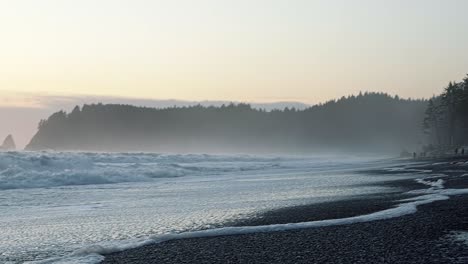 Beautiful-shot-of-the-coastline-of-the-famous-Ruby-Beach-near-Forks,-Washington-with-small-waves-crashing-and-leaving-sea-foam-behind,-and-large-cliffs-of-pine-trees-and-fog-on-a-warm-summer-evening