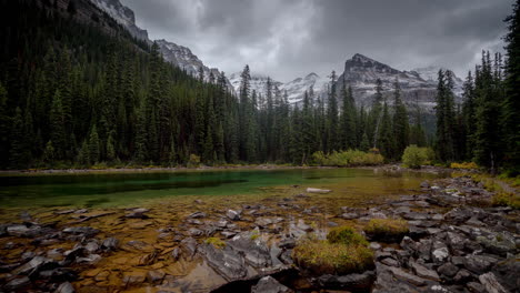 Time-Lapse,-Emerald-Alpine-Lake-Water-by-Conifer-Forest-and-Dark-Dramatic-Clouds-Above-Snow-Capped-Hills