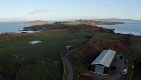 AERIAL---Beautiful-horizon,-a-warehouse-and-a-road,-Isle-of-Gigha,-Kintyre,-Scotland