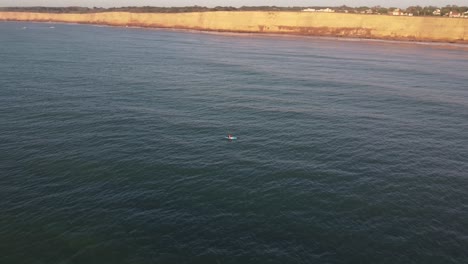 Aerial-orbiting-shot-of-a-person-with-kayak-rowing-in-wide-Atlantic-Ocean-front-to-cliffs-at-Acantilados-de-Mar-del-Plata-during-the-beautiful-sunset