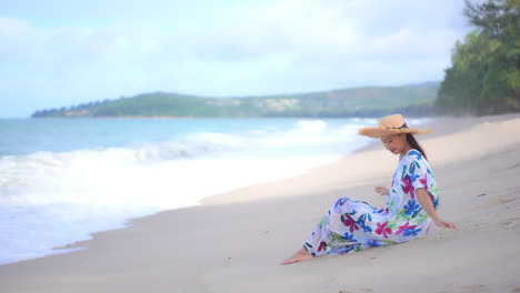 A-young-woman-holds-on-to-her-sun-hat-as-she-sits-on-the-beach-in-her-coverup