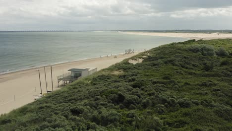 Aerial-shot-flying-over-green-dunes-and-past-a-beach-pavilion,-towards-the-sea-on-an-overcast-day