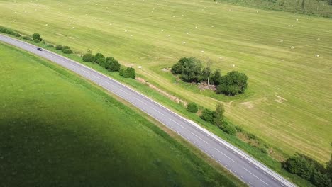 Aerial-of-an-asphalt-road-with-a-car-passing-by-next-to-a-large-empty-green-field-in-Estonia-during-summer