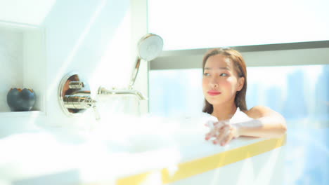 Beautiful-asian-woman-relaxing-in-bathtub-full-of-foam-in-bright-bathroom-on-sunny-morning,-full-frame