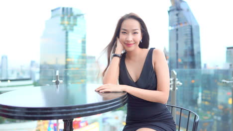 A-young-woman-in-a-little-back-dress-sits-at-a-table-on-a-rooftop-bar