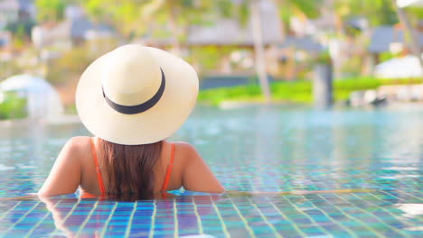 Close-up-of-a-woman-in-a-sunhat-and-relaxing-in-a-resort-swimming-pool-with-her-back-to-the-camera