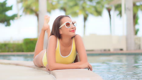 Lying-on-her-stomach-along-the-edge-of-a-posh-swimming-pool,-a-young,-fit-attractive-woman-smiles-as-she-looks-around