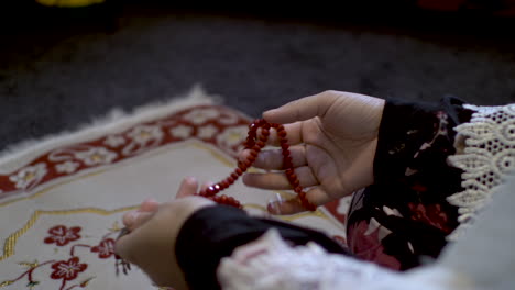 Over-The-Shoulder-View-Of-Female-Hands-Holding-Red-Prayer-Beads-Sitting-On-Prayer-Mat