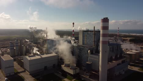 Steaming-chimney-vapour-emissions-floating-across-factory-refinery-warehouse-buildings-rising-aerial-shot