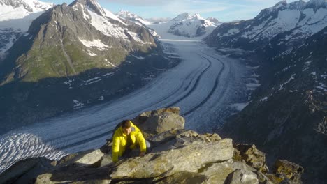 Young-man-in-yellow-jacket-on-rock-formation,-hiking-on-rocks,-overlooking-epic-Aletsch-Glacier,-biggest-glacier-in-the-Alps