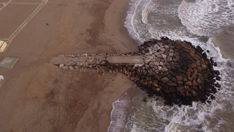 Kreisender-Schuss-Der-Wasserwellen,-Die-Die-Felsen-Zerschmettern,-Wo-Leute-An-Einem-Milden-Klimatag-In-Mar-Del-Plata,-Argentinien,-über-Den-Wellenbrecher-Am-Strand-Gehen