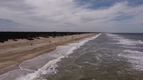 Drone-flying-over-ocean-waves-with-cars-driving-on-sandy-beach,-Mar-de-las-Pampas-in-Argentina
