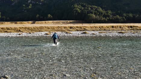 Woman-with-backpack-walking-crossing-flowing-river-at-hike-in-New-Zealand-during-sunny-day---rear-view