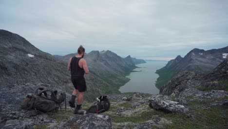 Male-Hiker-Walks-Toward-Alaskan-Malamute-Dog-Relaxing-On-The-Ground-At-Anderdalen-National-Park,-Overlooking-Fjord-In-Senja,-Norway