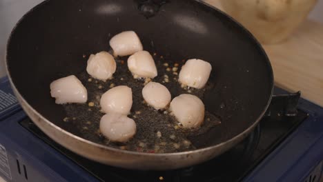 Home-cooking-on-butane-gas-stove,-chef-swirling-the-hot-oil-and-garlic-in-the-pan-in-slow-motion,-making-sure-all-scallops-are-cooked-evenly,-close-up-shot