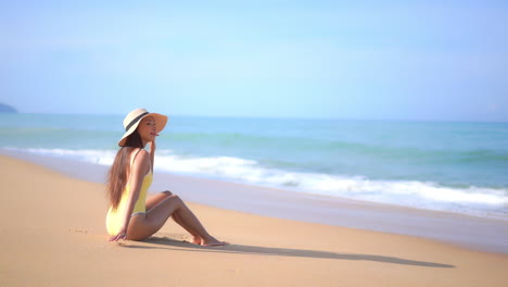 A-young-woman-in-a-yellow-bathing-suit-sits-on-a-sandy-beach-enjoying-the-scenery