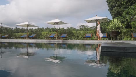 Fancy-blond-woman-in-swimwear-cover-up-dress-walking-along-pool-at-Bali-resort,-reflection-on-water