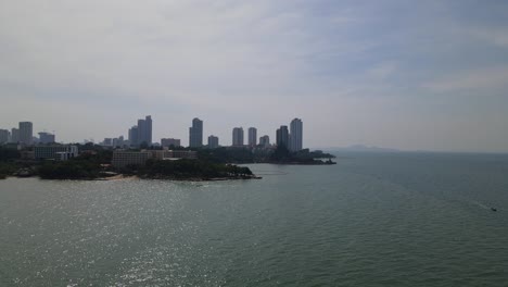 Reverse-aerial-footage-of-the-Sanctuary-of-Truth-in-a-silhouette-and-a-beachfront-with-high-rise-condominiums-in-the-background-while-a-boat-speeding-on-the-right,-Pattaya,-Chonburi,-Thailand