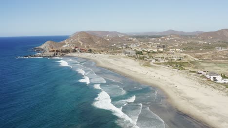 Wide-angle-slow-motion-shot-of-the-Los-Cerritos-beach-water-waves-crashing-to-the-sand-in-a-mountain-range-near-a-coast-or-a-town-on-a-sunny-day-in-Mexico