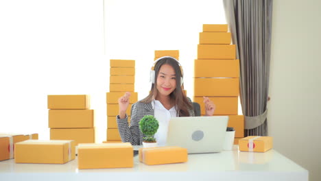 While-surrounded-by-packages-ready-for-shipping,-a-young-woman-seated-in-front-of-a-laptop-does-a-happy-success-dance