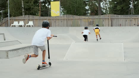 Three-friends-boys-riding-ride-kick-bike-kickbikes-scooter-electric-scooters-skateboard-skate-park-concrete-ramp-jump-jumps-of-edge-flying-tricks-kid-stunt-professional-summer-camp-cool-evening-boy